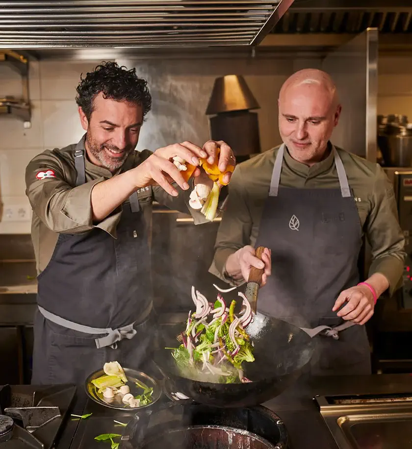 Two chefs cooking together in a professional kitchen, one adding ingredients to a wok while the other tosses vegetables in the pan.